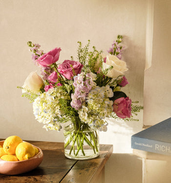 Bouquet of flowers in a vase on a wooden surface with a book and bowl of lemons in the background.