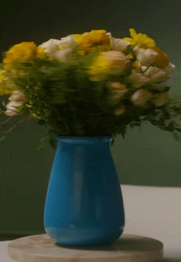 Animated 360° spin of a yellow and white floral bouquet in a glossy blue vase on a light marble turntable, against a dark olive-green backdrop.